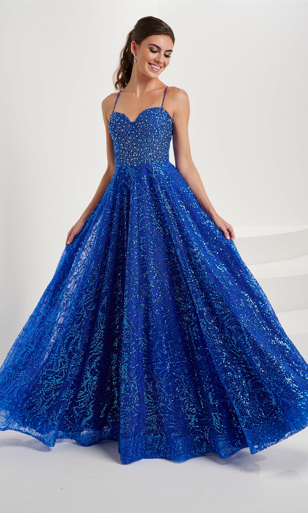 A woman wears the Tiffany Beaded-Bodice Glitter Tulle Prom Dress 16088, featuring a sleeveless royal blue beaded bodice and a full, floor-length glitter tulle skirt, standing against a white background.